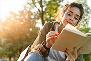 Young woman sitting outdoors, reading a book with a smile. Sunlight filters through the trees, creating a warm, serene atmosphere. Relaxed and content.